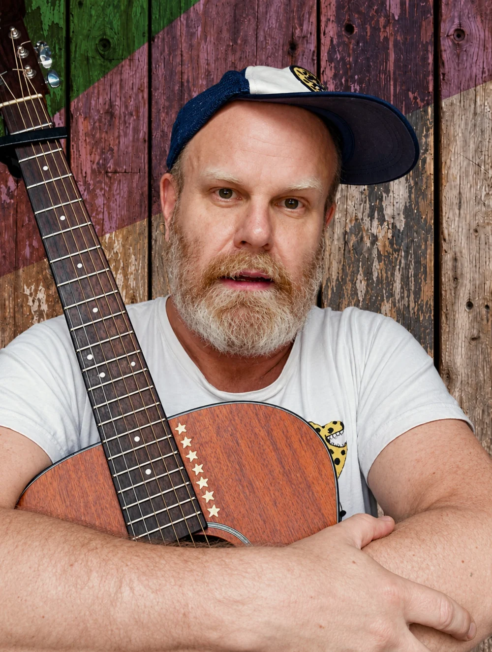 Jay Bones facing the camera with his acoustic guitar against a painted wooden background